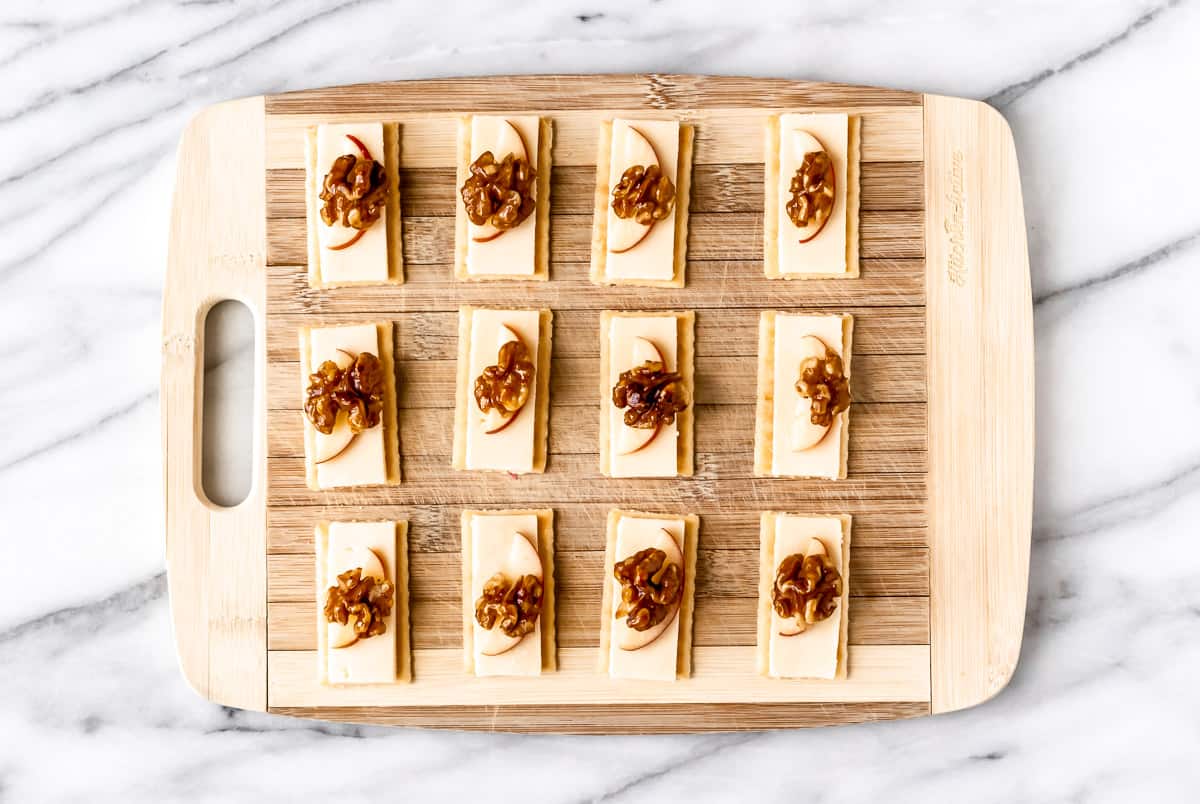 Crackers on a cutting board topped with cheese, apples and honey walnuts.