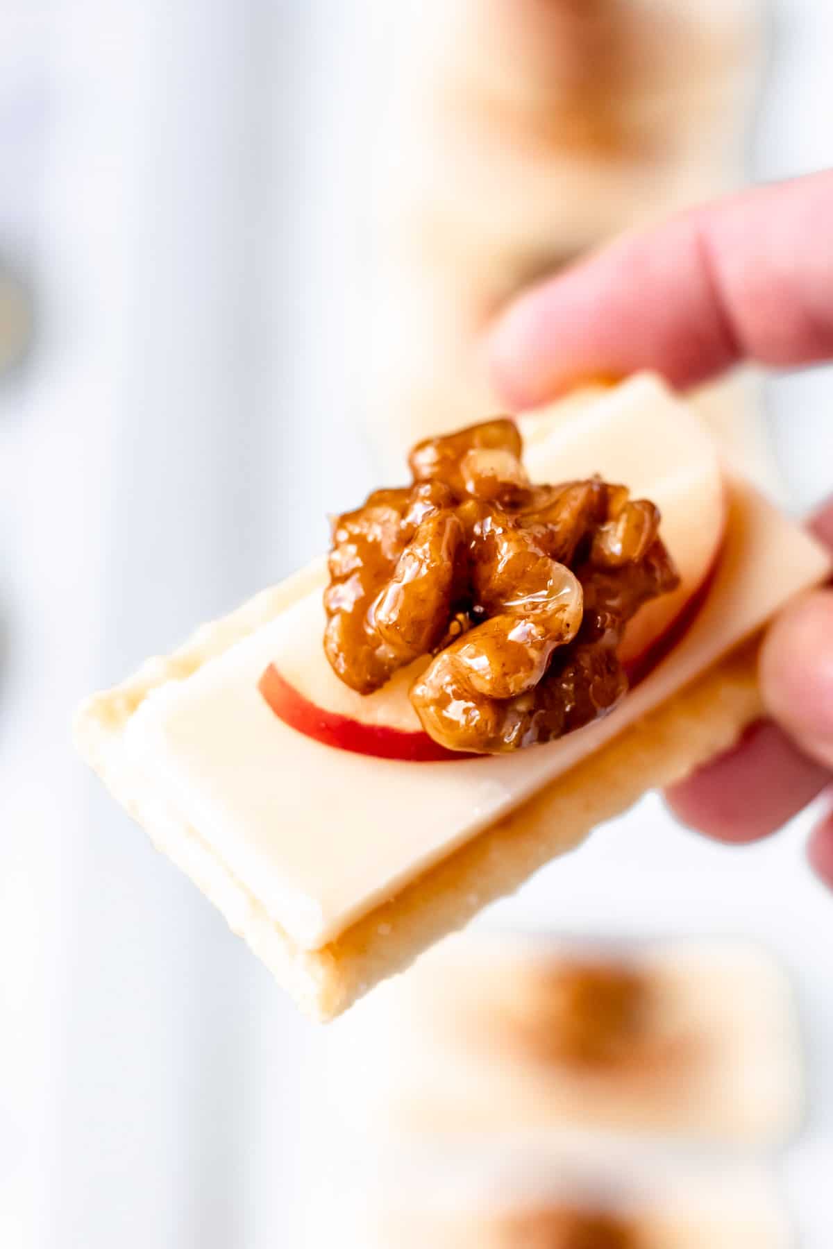 A hand holding up a single cracker with cheddar cheese, apples and walnuts on it.