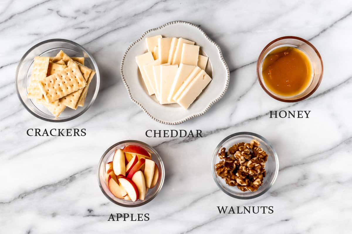 Ingredients to make cheese and crackers with apples and honey walnuts on a marble background with text overlay.