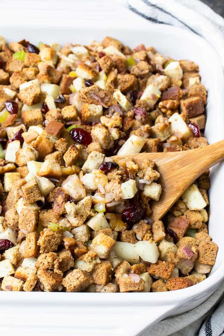 close up of apple cranberry walnut stuffing in a white casserole dish with a wooden spoon lifting some out