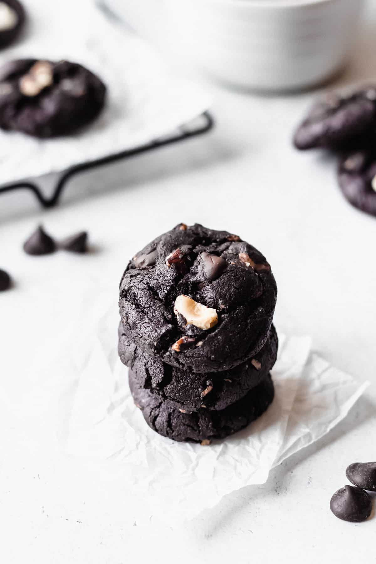 A stack of three dark chocolate hazelnut cookies on white paper with more cookies around them