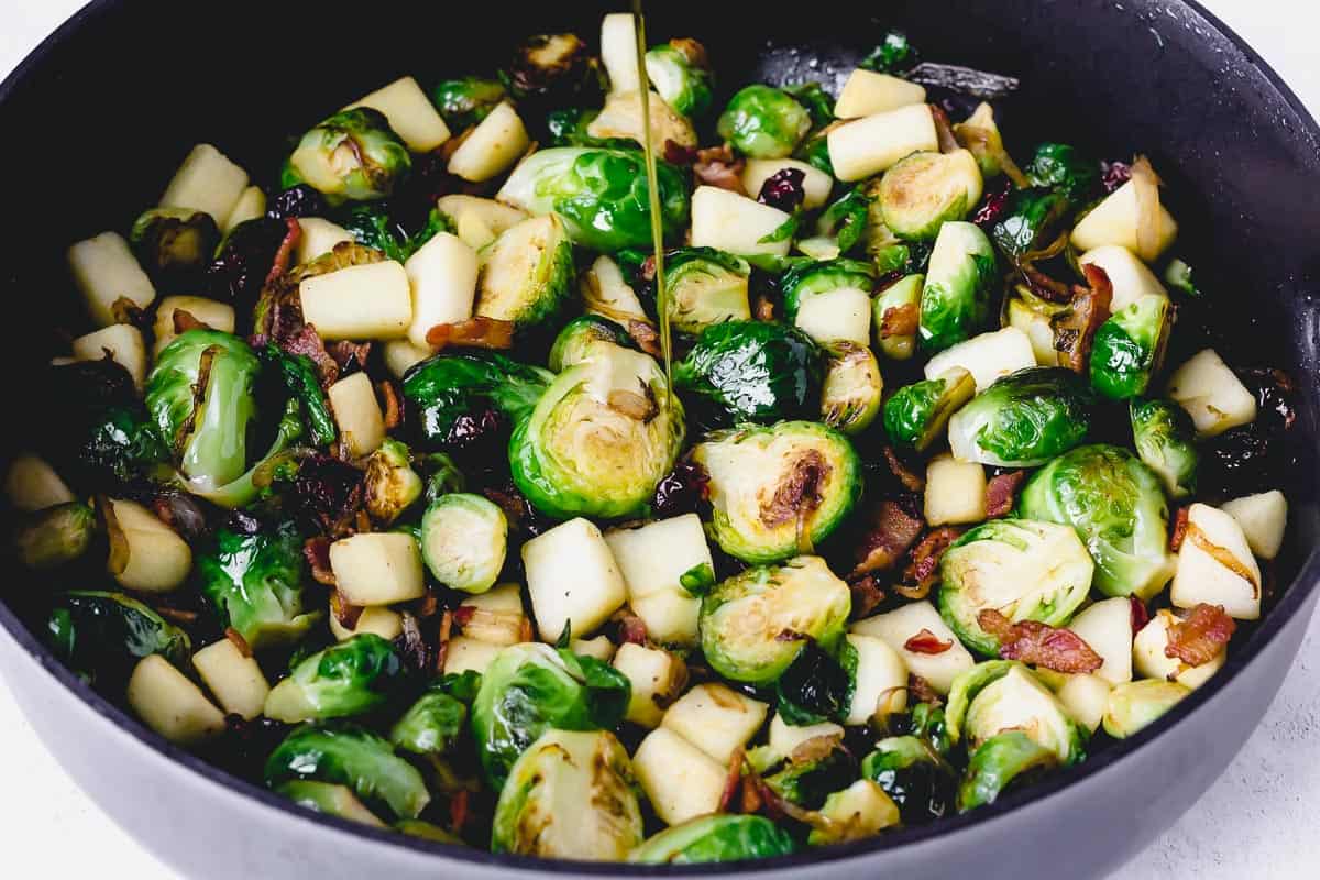 Maple syrup being poured onto brussels sprouts and fruit in a black skillet