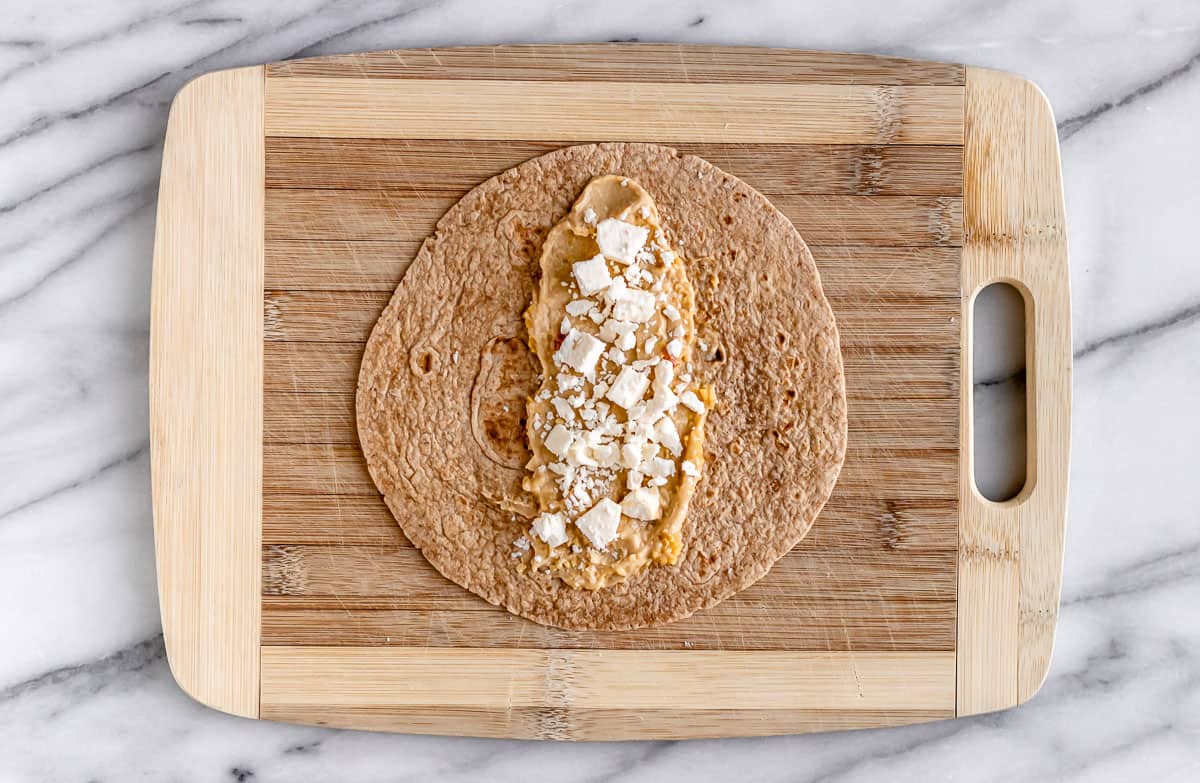 A tortilla on a cutting board topped with hummus and feta cheese.