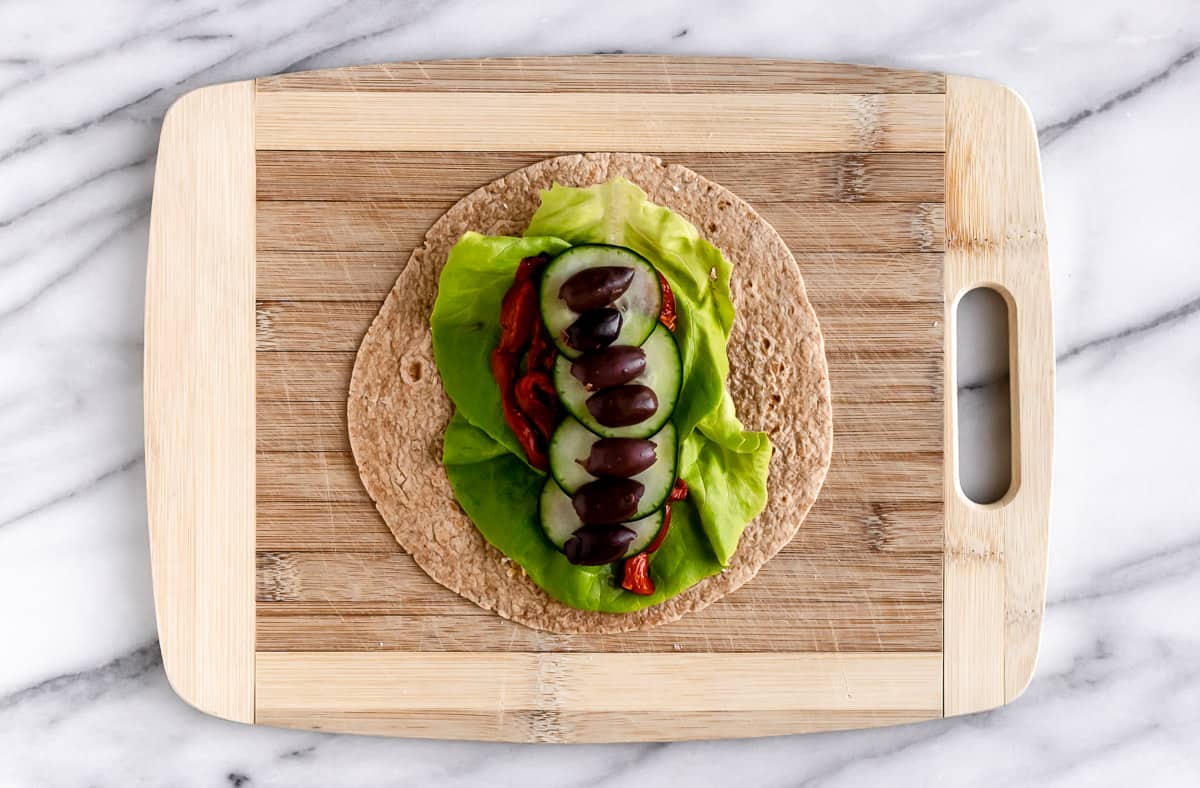 A tortilla on a cutting board topped with lettuce, peppers, cucumber and olives.