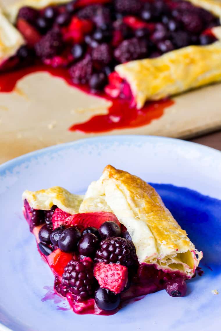 A pieces of Mixed Berry Puff Pastry Tart on a blue plate with the rest of the tart in the background on a wood backdrop