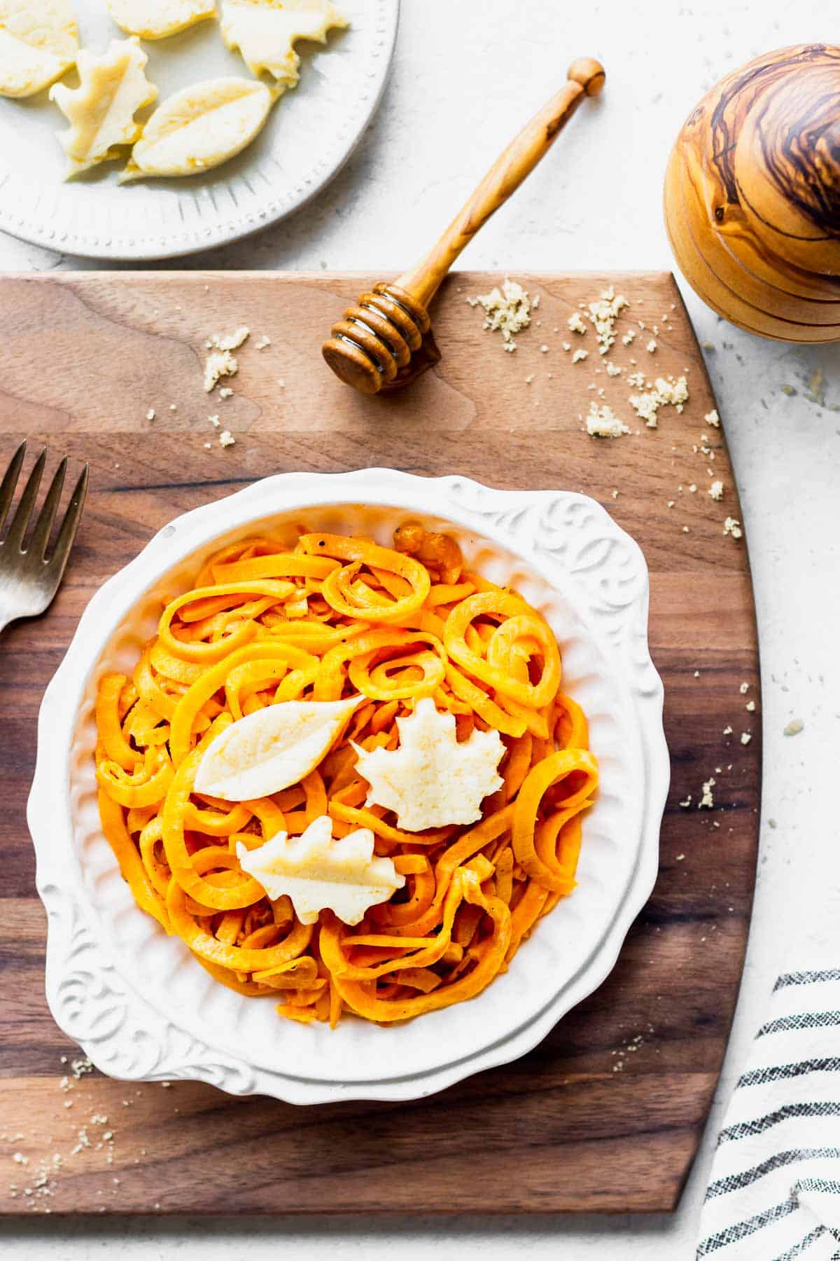 A bowl of spiralized sweet potato on a wood board with a honey dipper, honey pot, and brown sugar around it and a small plate of butter above it.