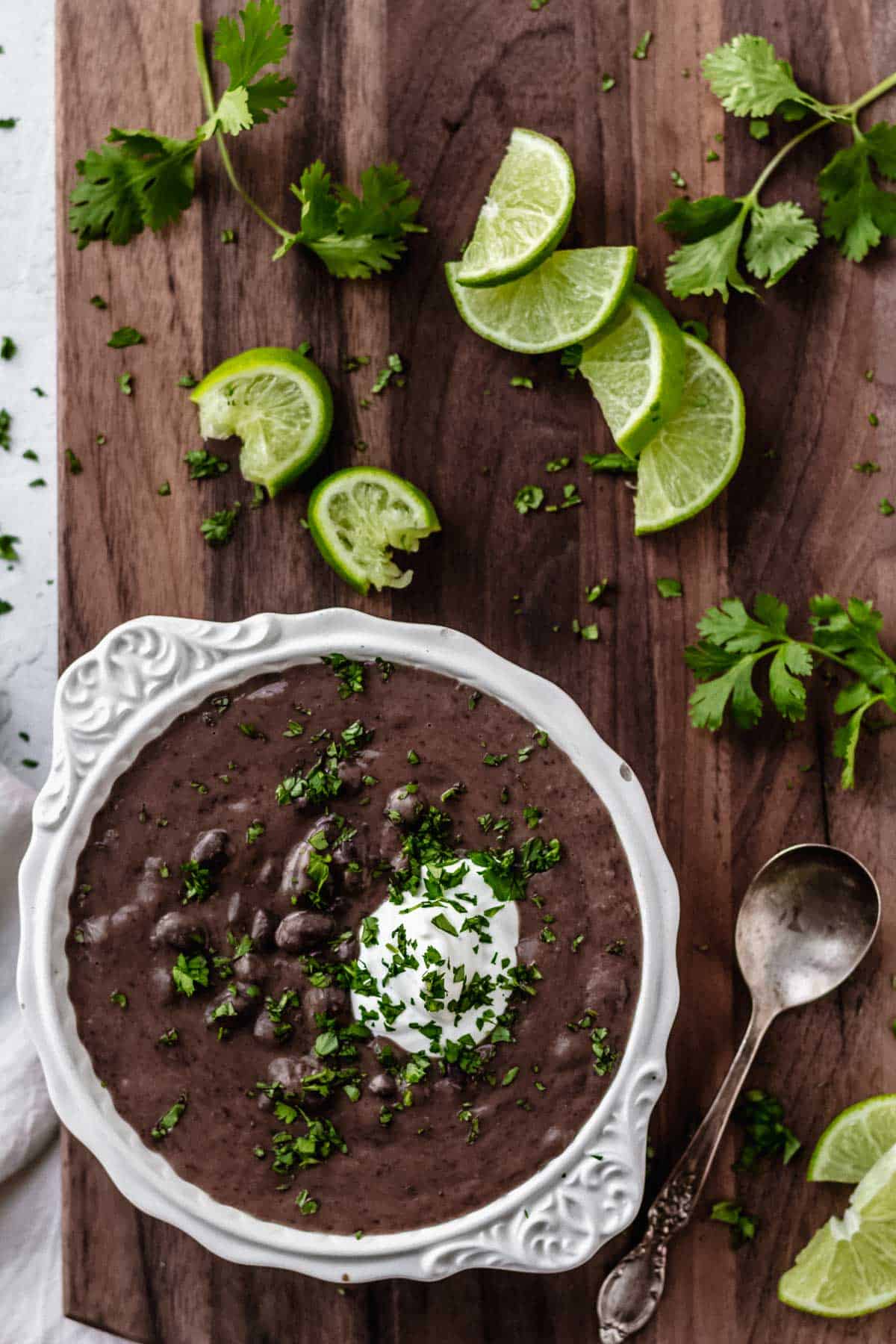 Overhead of black bean and lime soup in a white bowl on a wood board with lime slices, a spoon, and cilantro