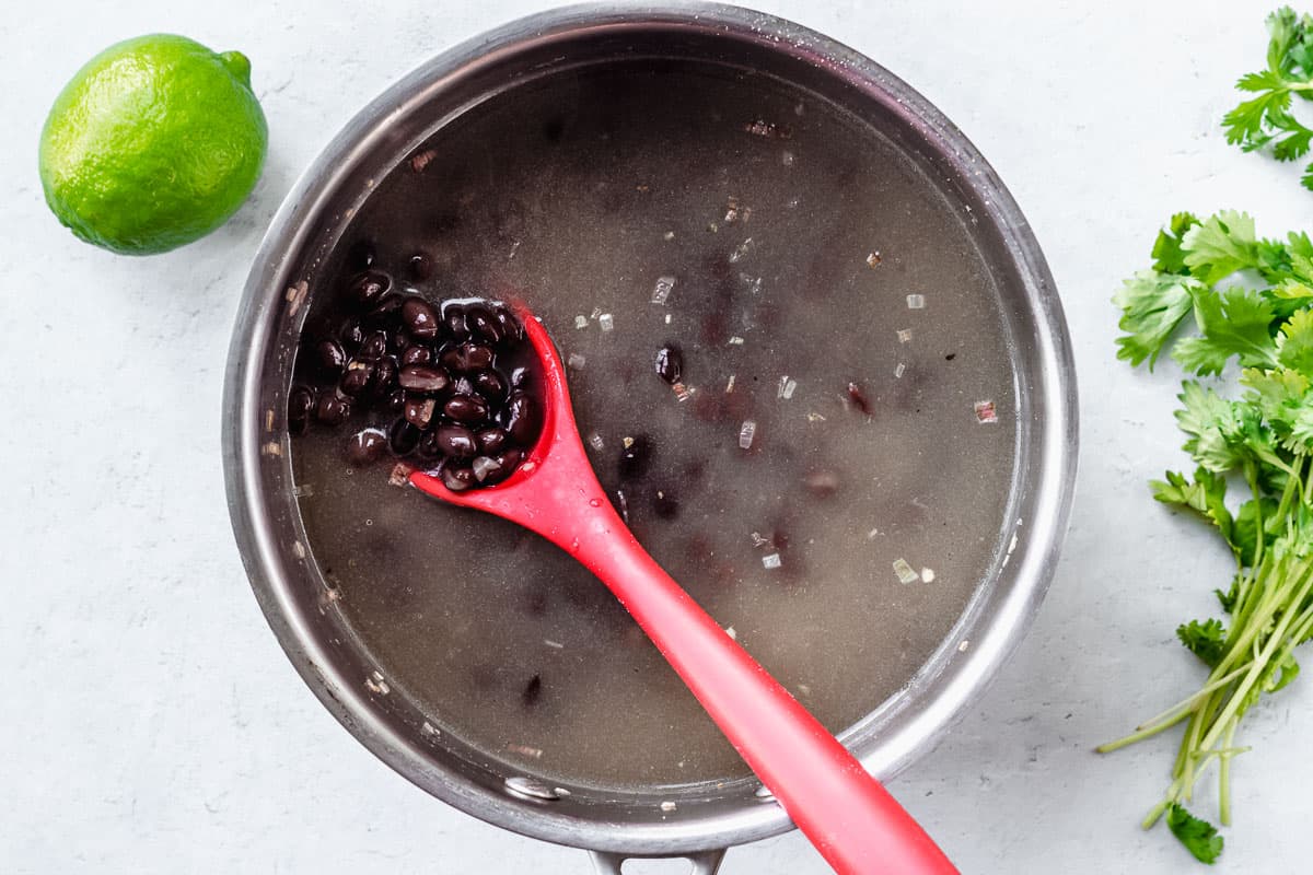 Black beans cooking in broth in a pot over a white background with a lime and cilantro around it