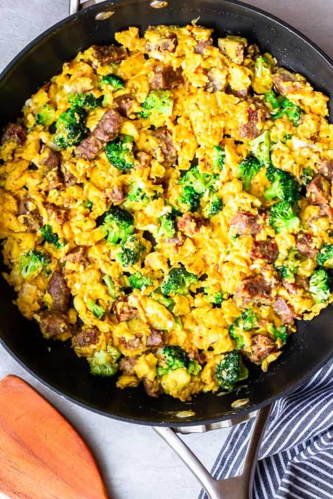 Scrambled eggs and sausage with cheese and broccoli in a black skillet with a blue and white towel and wood turner around it