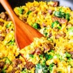 close up of scrambled eggs with sausage, cheese, and broccoli in a black skillet with a wood turner lifting some up