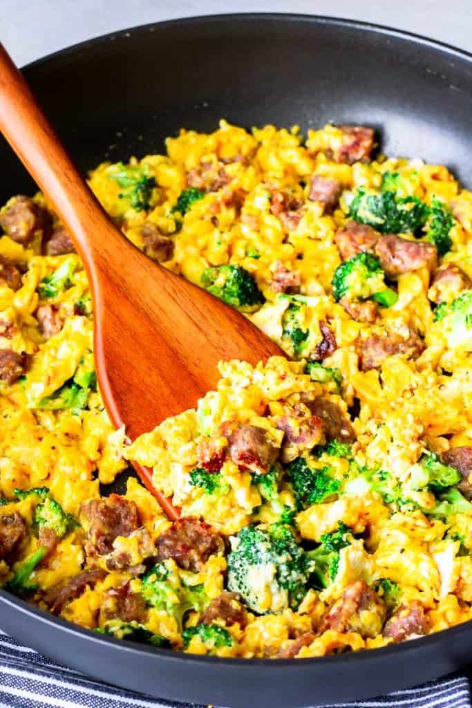 close up of scrambled eggs with sausage, cheese, and broccoli in a black skillet with a wood turner lifting some up
