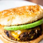 A southwestern black bean burger on a toasted bun with melted cheese and avocado on a white plate with a wood background