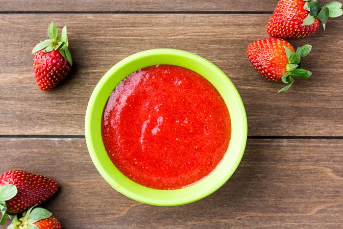 A green bowl of Strawberry Puree with 5 Strawberries Around It on a wood background