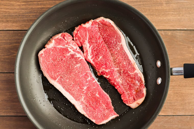 2 steaks cooking in a black skillet over a wood background