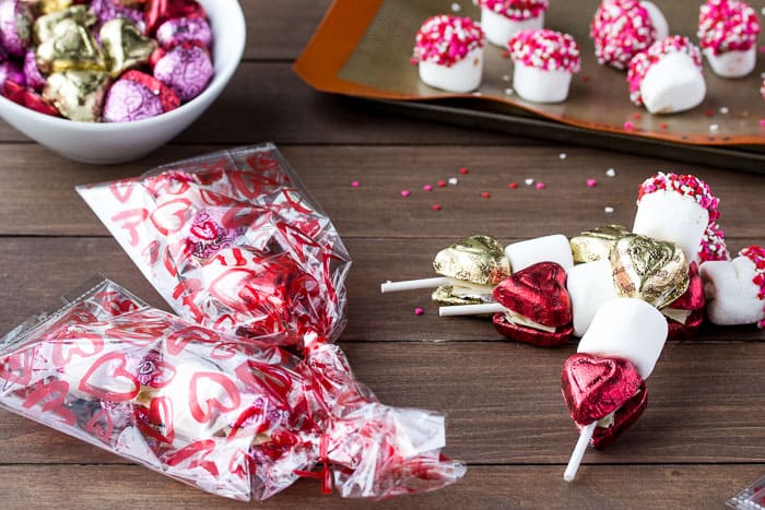2 Valentine's Day Candy Kabobs in Gift Bags, 2 not in gift bags with extra candy and chocolate dipped marshmallows in the background all on a wood surface with extra sprinkles scattered around