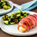 bacon wrapped chicken with brussels sprouts on a white plate with a second plate in the background, a blue napkin, and a fork over a wood background
