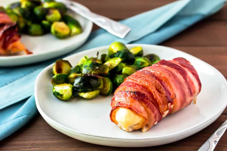 bacon wrapped chicken with brussels sprouts on a white plate with a second plate in the background, a blue napkin, and a fork over a wood background