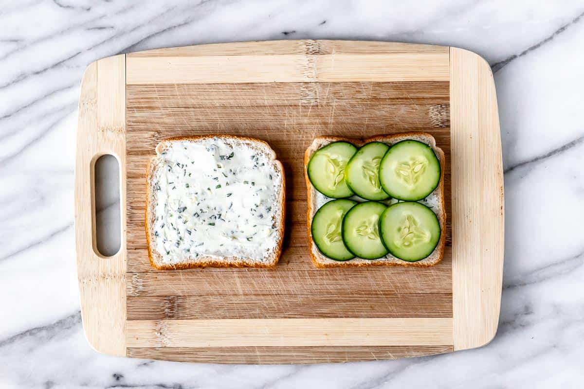 Two slices of bread on a cutting board with cream cheese on one and cream cheese and slices of cucumber on the other.