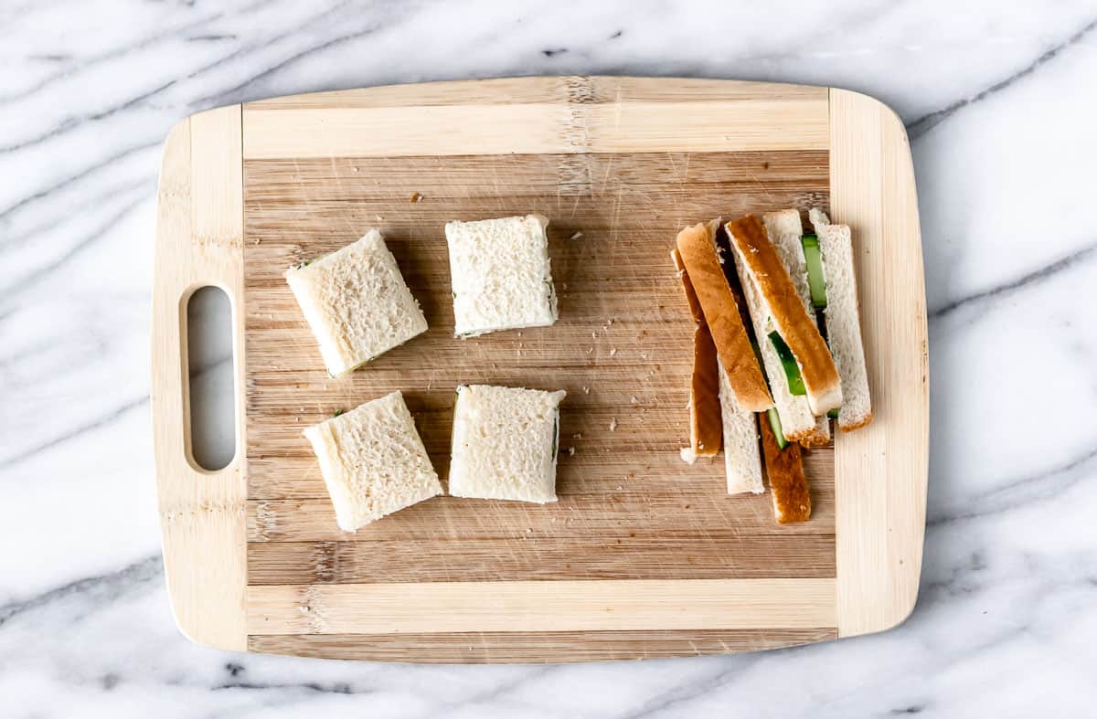 Four cucumber tea sandwiches on a cutting board with the crust of the bread off to the side.