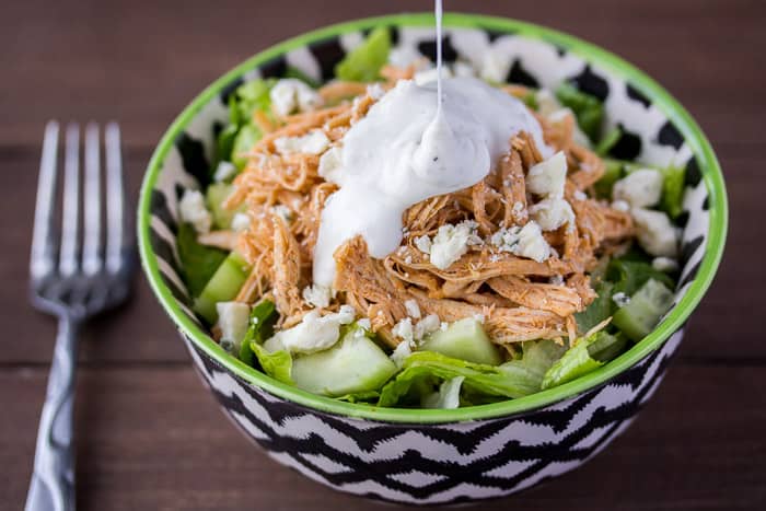 Ranch dressing being poured onto a buffalo chicken salad in a black and white bowl with a fork next to it on a wood background