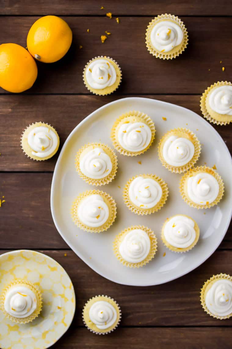Overhead view of Mini lemon cupcakes on a white round tray with cupcakes scattered around it and 2 lemons all over a wood background