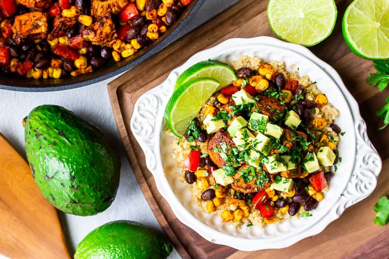 Overhead of A white bowl filled with quinoa, chicken, black beans, corn, red pepper, avocado, and lime slices on a wood board with 2 lime halves, and part of a skillet in the background