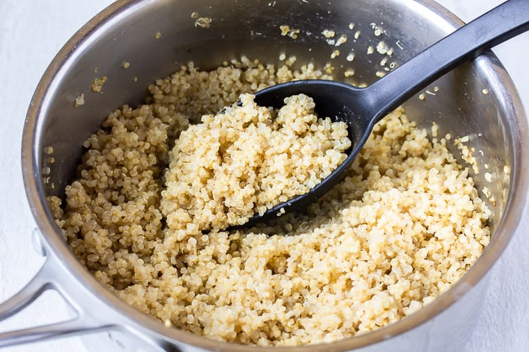 A silver pot with cooked quinoa and a black spoon in it over a white background