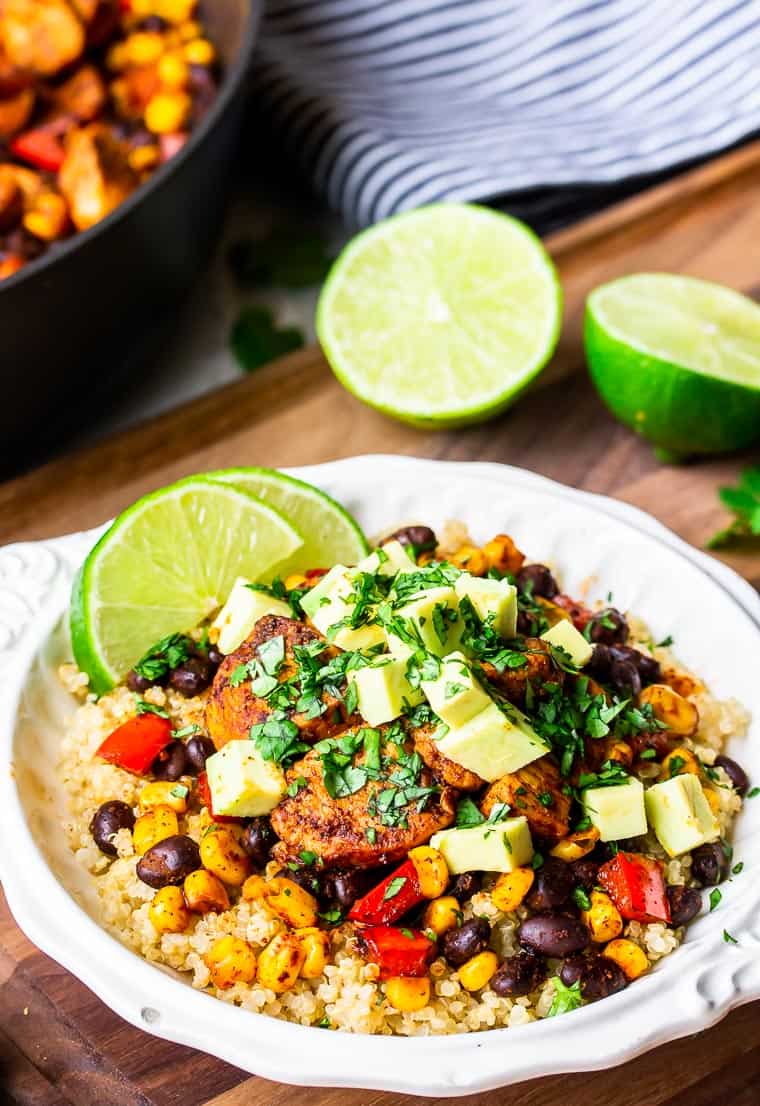 A white bowl filled with quinoa, chicken, black beans, corn, red pepper, avocado, and lime slices on a wood board with 2 lime halves, a blue and white towel and part of a skillet in the background