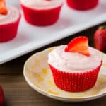 A single strawberry cupcake on a light orange plate with a white tray of more cupcakes in the background and strawberries scattered around them.