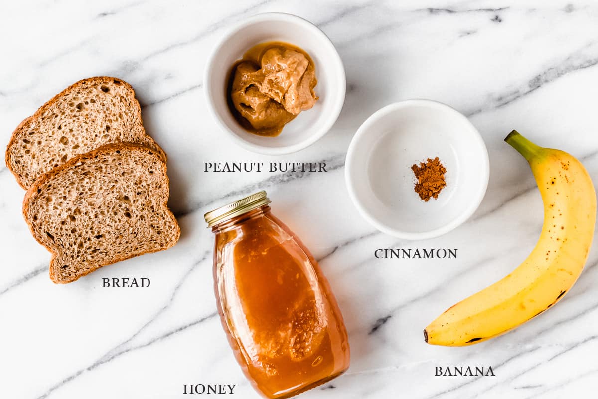 Ingredients to make banana toast with peanut butter, honey and cinnamon on a marble background with labels