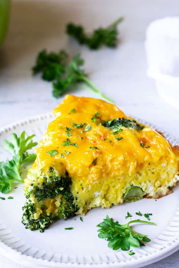 Close up of a slice of broccoli cheddar frittata on a white plate with parsley in the backrground