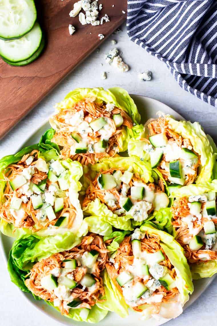 Overhead of a Buffalo Chicken Lettuce Wraps on a white plate with a cutting board, cucumber slices, blue cheese, and a blue and white towel in the background.