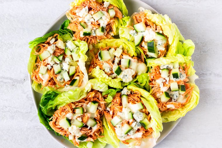 Buffalo Chicken filled lettuce cups topped with blue cheese, ranch dressing and cucumber on a white plate over a white background