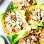 Overhead of half a plate of Buffalo Chicken Lettuce Cups on a white background with a cutting board, cucumber slices and blue cheese around it.