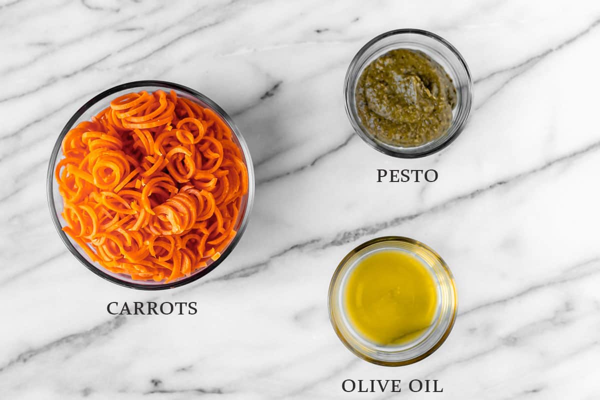 Ingredients needed to make pesto carrot noodles on a marble background with text overlay.