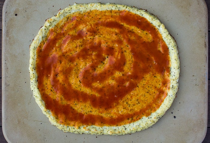 Buffalo Sauce Spread Onto The cauliflower Pizza Crust on a pizza stone
