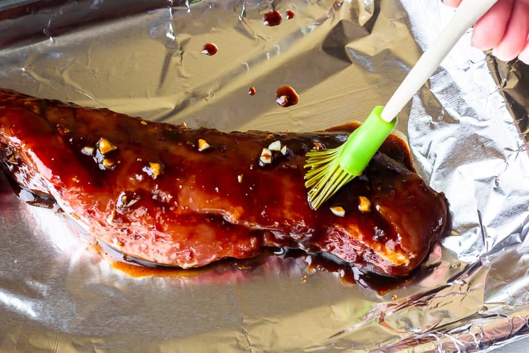Basting a pork tenderloin with Asian sauce and a green silicone spoon on a foil-line baking sheet
