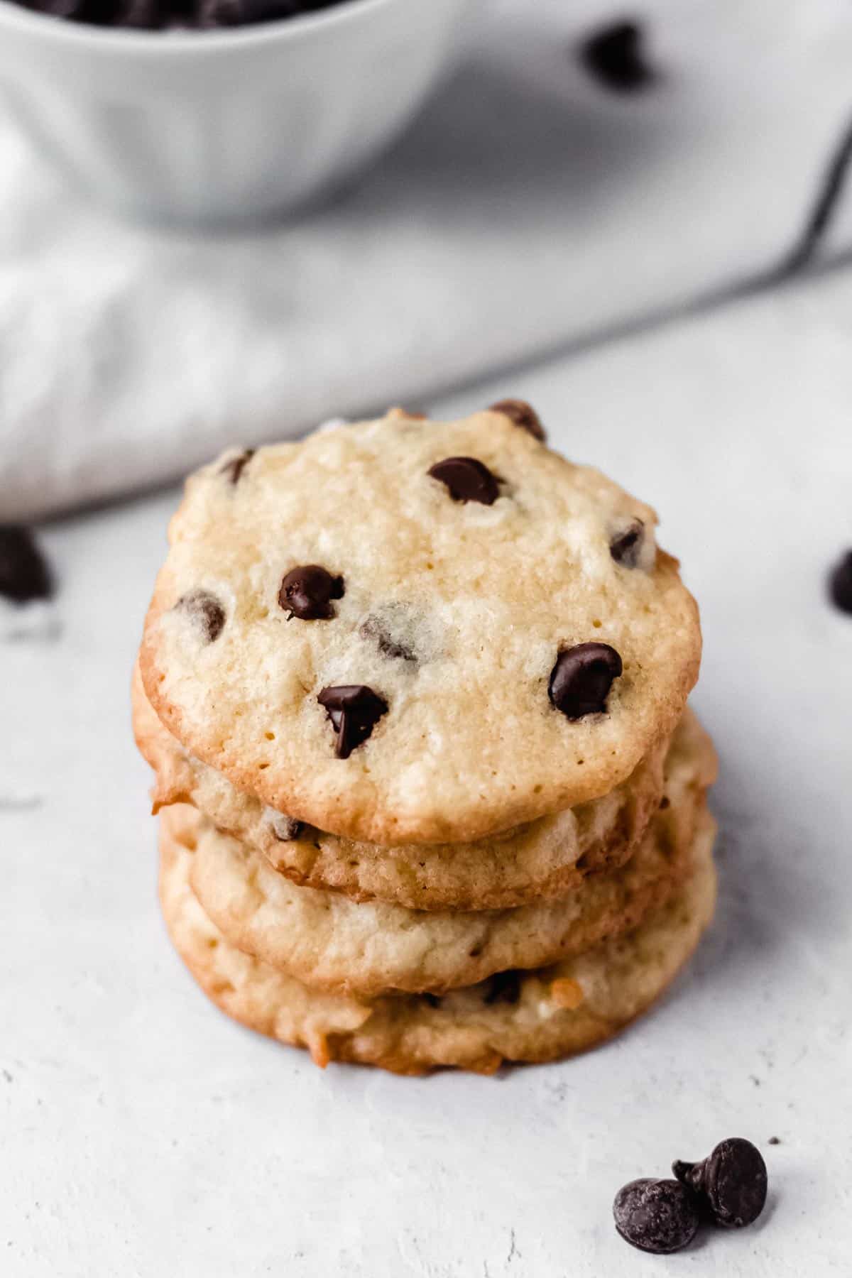 Stack of 5 coconut chocolate chip cookies on a white background with chocolate chips, a white towel and a bowl in the background