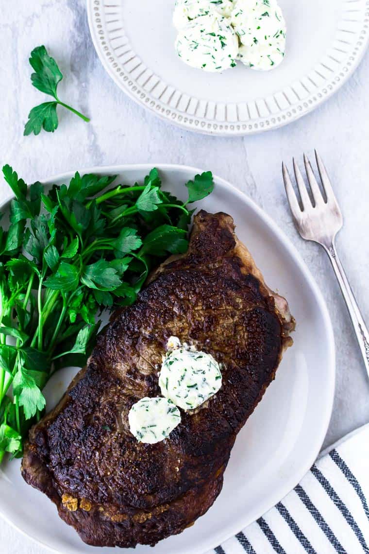 A white plate with a steak topped with 2 pats of garlic herb steak butter with a bunch of parsley next to it and a smaller plate of extra butter, a fork and black and white napkin all around it