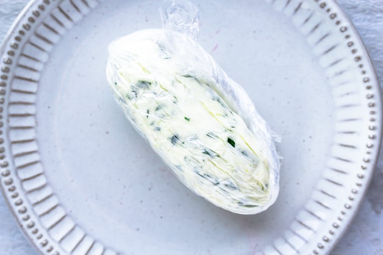 compound butter rolled into a log and wrapped in plastic wrap on a white plate over a white background