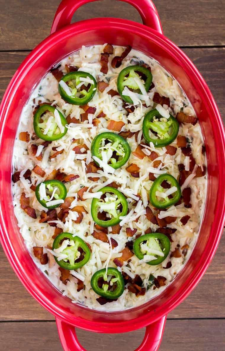Overhead View of a Unbaked Jalapeno Popper Casserole Topped with Extra Cheese, Bacon, and Jalapeno Pepper Slices in a red casserole dish on a wood background.
