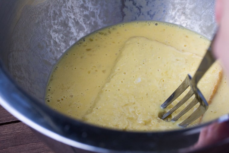 A Piece of Sourdough Bread Being Dipped into the Egg Mixture in a silver bowl with a fork