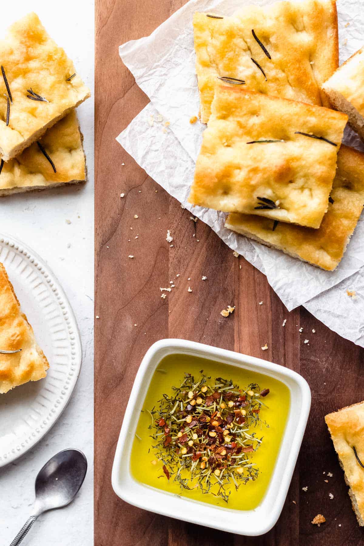 Bread dipping oil and pieces of focaccia bread on a wood cutting board with extra pieces and a small plate and spoon off to the side