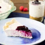 A Slice of Raspberry No-Bake Cheesecake on a Blue Plate with the Pie and a Mason Jar in the Background