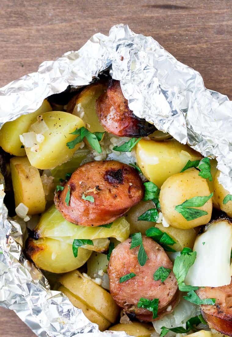 Close up of sausage, cabbage and potatoes topped with parsley in a foil packet over a wood background