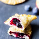 2 halves of a mini blueberry hand pie broken open with blueberry filling inside on a piece of slate with more pies and fresh blueberries around it