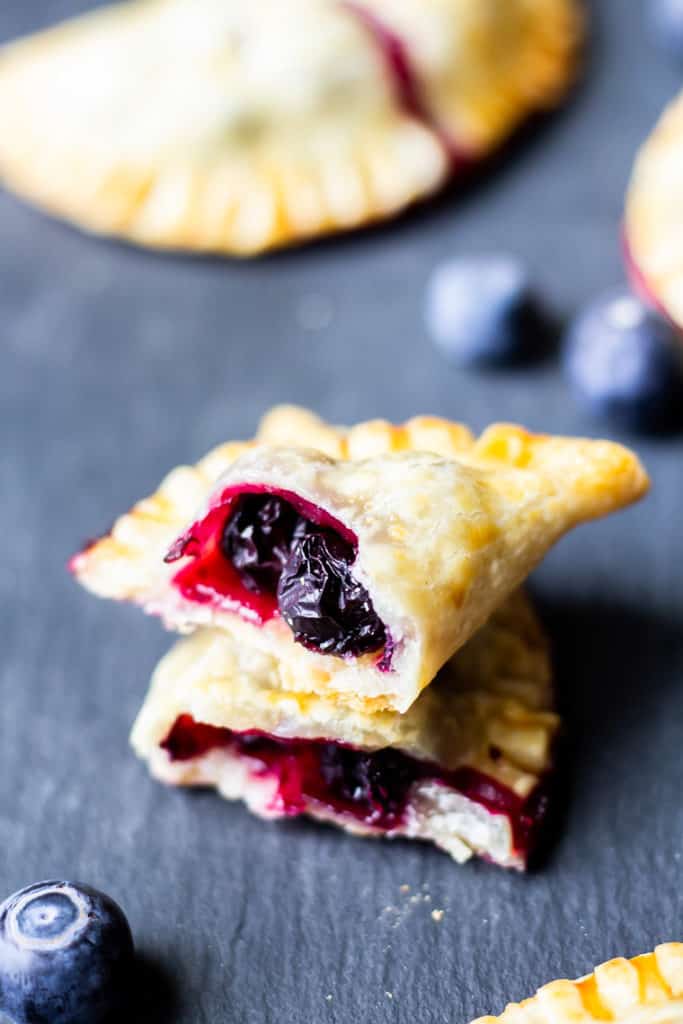 2 halves of a mini blueberry hand pie broken open with blueberry filling inside on a piece of slate with more pies and fresh blueberries around it