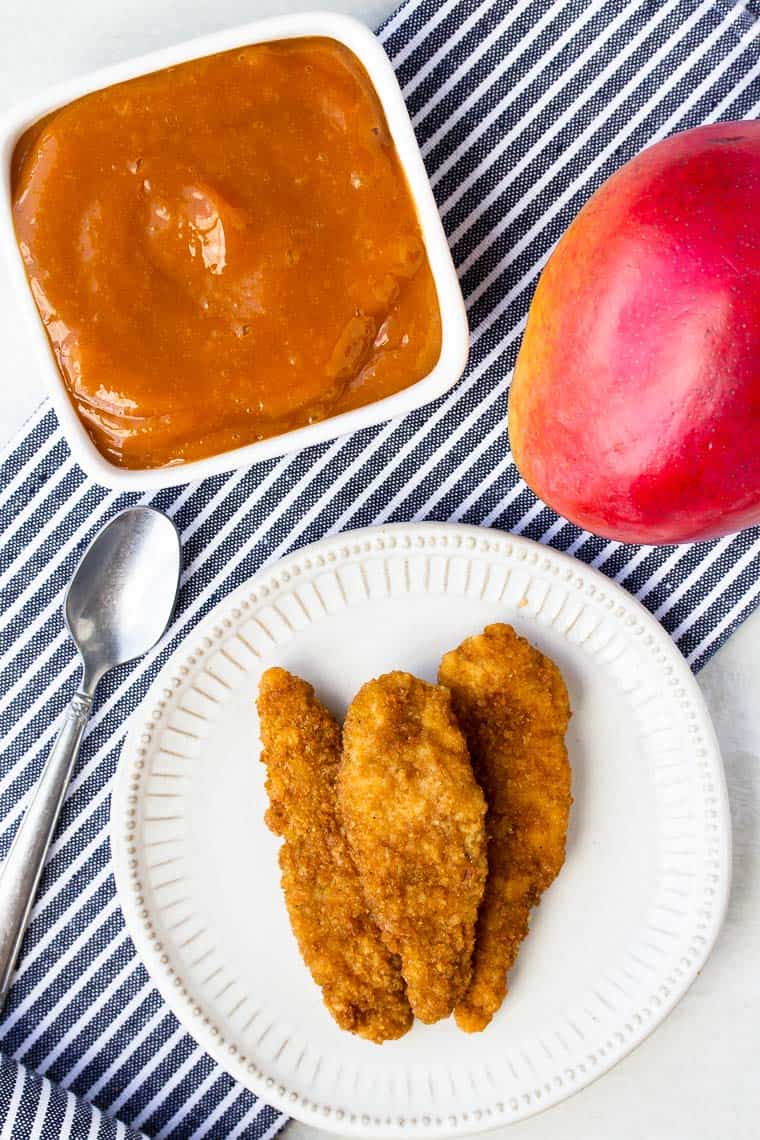 Overhead view of Mango Barbecue Sauce in a white square dish, a small spoon, a mango, and chicken tender on a white plate over a blue and white striped napkin