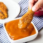 A hand dipping a chicken tender into a square dish of Mango Barbecue Sauce with a blue and white napkin and small plate of more chicken in the background