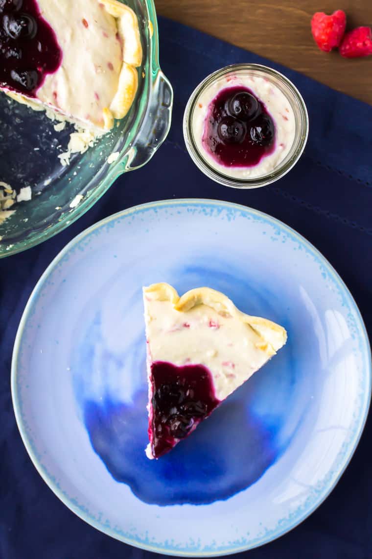 Overhead of a no bake raspberry cheesecake slice on a blue plate and in a mason jar with part of the pie showing and 2 raspberries in the background all over a wood background